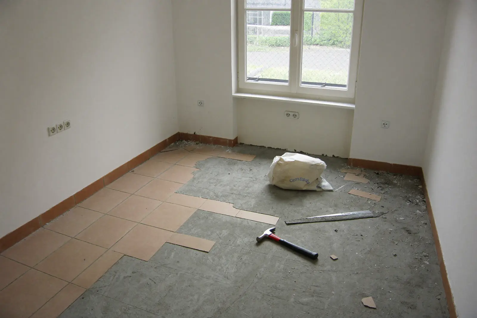 Lege woonkamer in een jaren dertig woning waar de helft van de oude tegelvloer al is verwijderd en de grijze dekvloer zichtbaar is