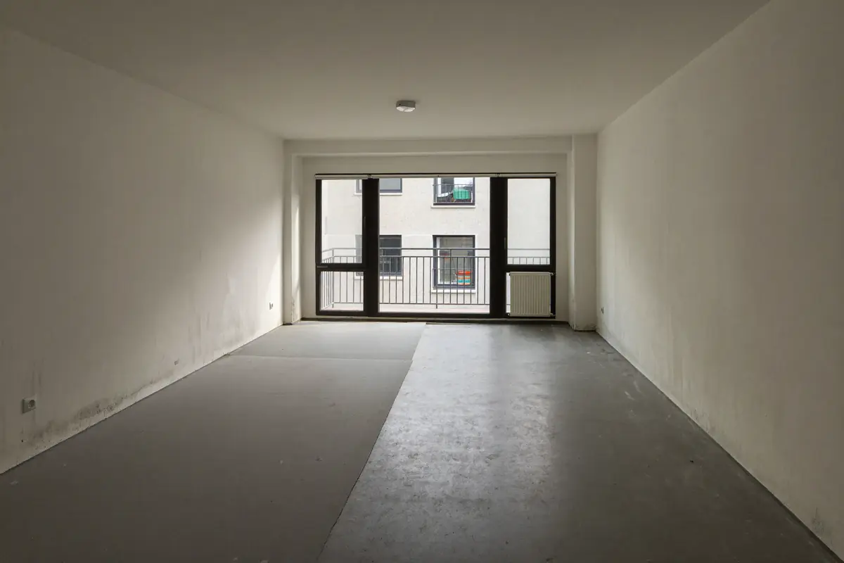 Lege woonkamer in een appartement met een kale, schoongeveegde betonvloer en grote ramen naar het balkon