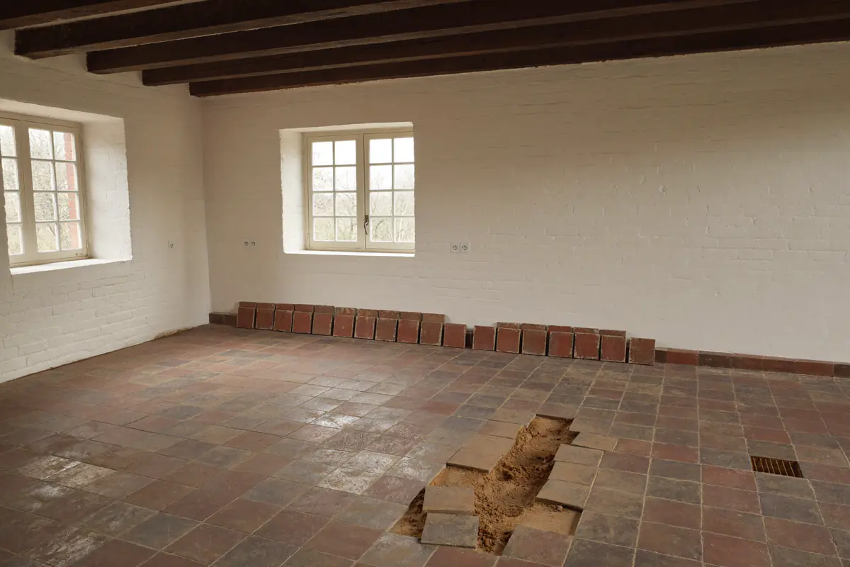 Boerderij-interieur met rode plavuizen in een zandbed, waarvan een deel al is gelicht en langs de muur is gestapeld